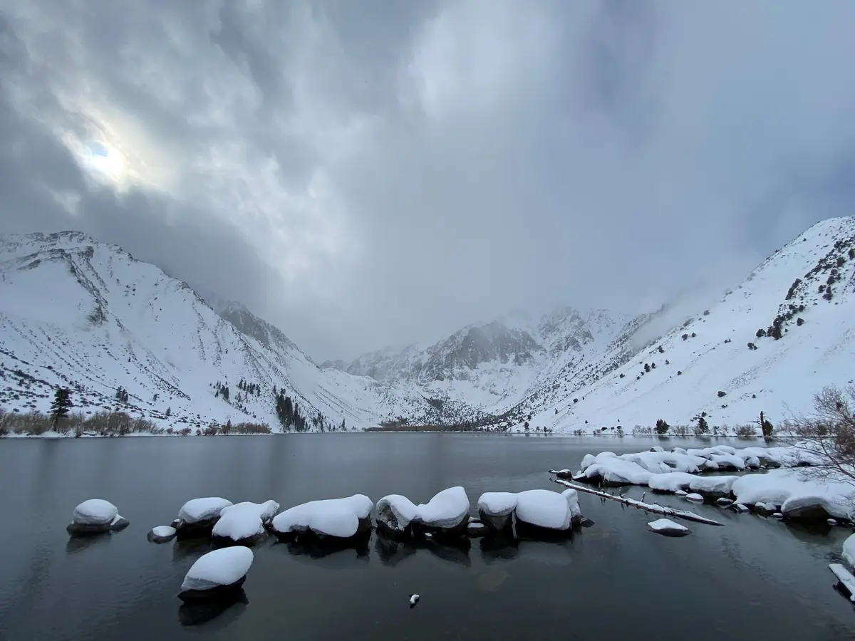 Snow-capped boulders in the dark water of Convict Lake, below snowy peaks and moody winter clouds