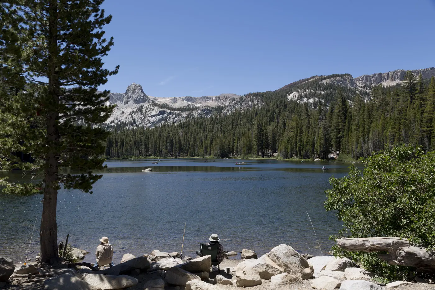Two anglers fishing from the rocky shore of Lake Mary, with boats on the water and the granite spire of Crystal Crag rising above the pine forest
