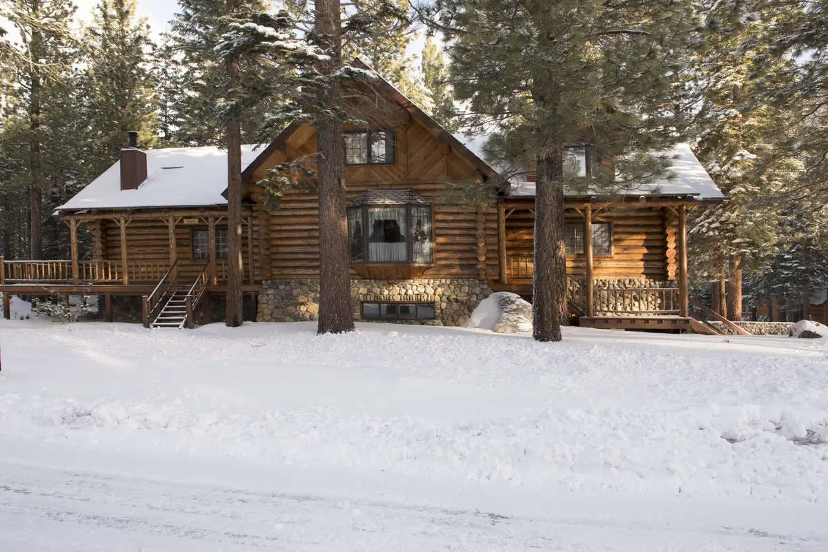 A rustic log cabin with a stone foundation and wraparound deck, set among tall pines in fresh snow in Mammoth Lakes
