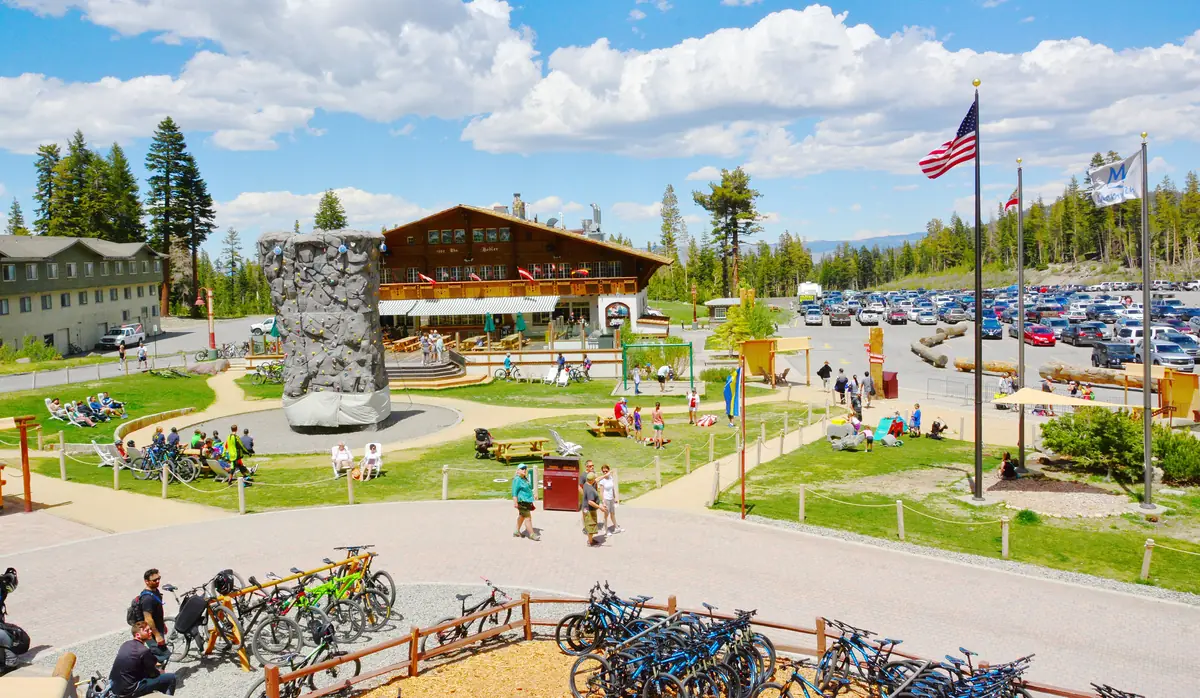 The Mammoth Mountain Main Lodge area in summer, with racks of mountain bikes, a climbing wall, green lawns and visitors under puffy clouds