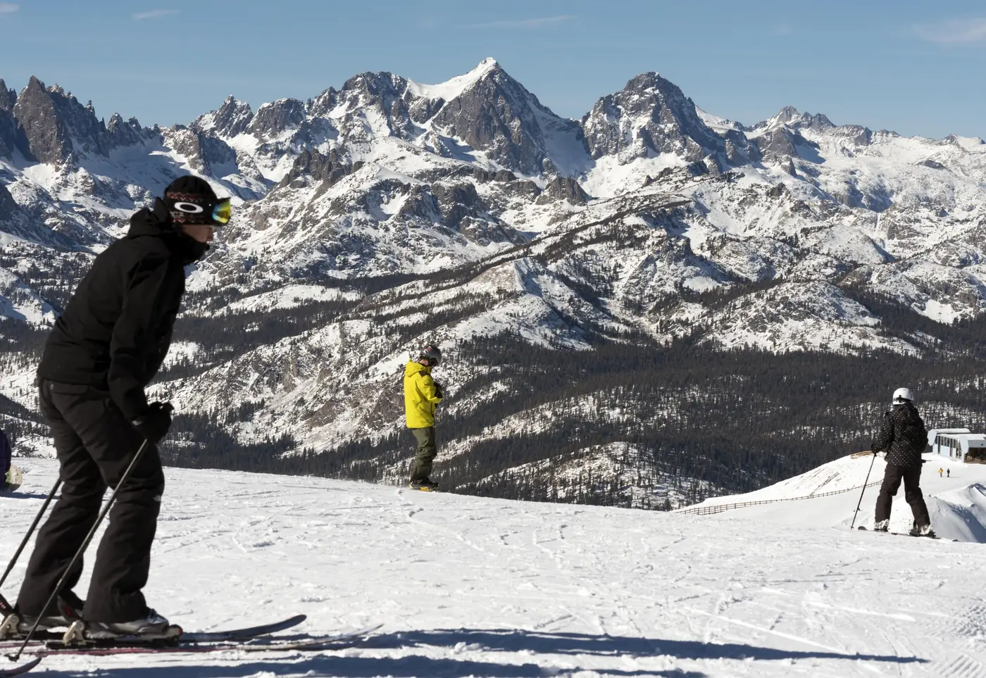 Skiers pausing on a groomed run at the top of Mammoth Mountain, with the jagged snow-covered Minarets and Ritter Range under a clear blue sky